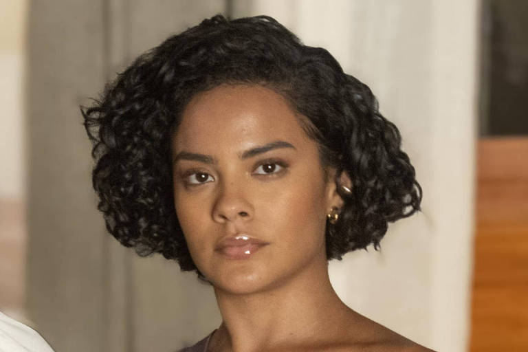 A imagem mostra duas mulheres em pé, uma ao lado da outra. A mulher à esquerda tem cabelo loiro e usa uma blusa branca com um colar de pérolas. A mulher à direita tem cabelo curto e cacheado, vestindo uma blusa de um ombro só na cor roxa. O fundo é composto por paredes claras e janelas de madeira.