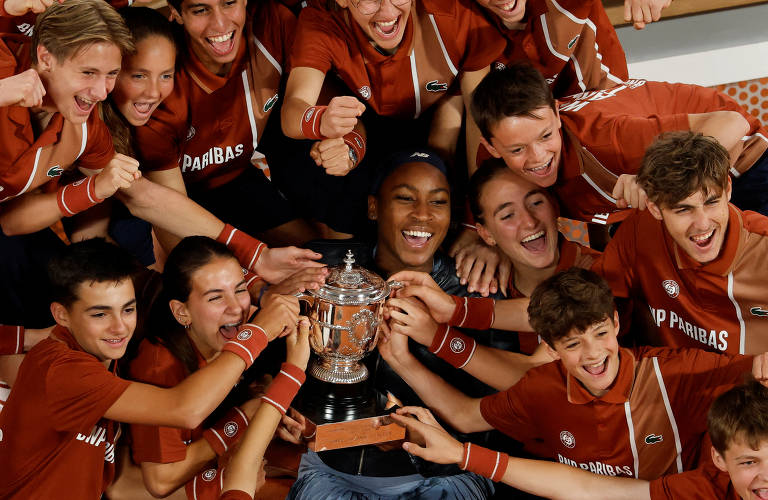 Um grupo de jovens atletas, todos vestindo camisetas marrons com detalhes em branco, estão reunidos em torno de uma troféu de tênis. Eles estão sorrindo e celebrando, com alguns segurando o troféu e outros levantando as mãos em sinal de alegria. A atmosfera é de comemoração e união, com todos parecendo felizes e animados.