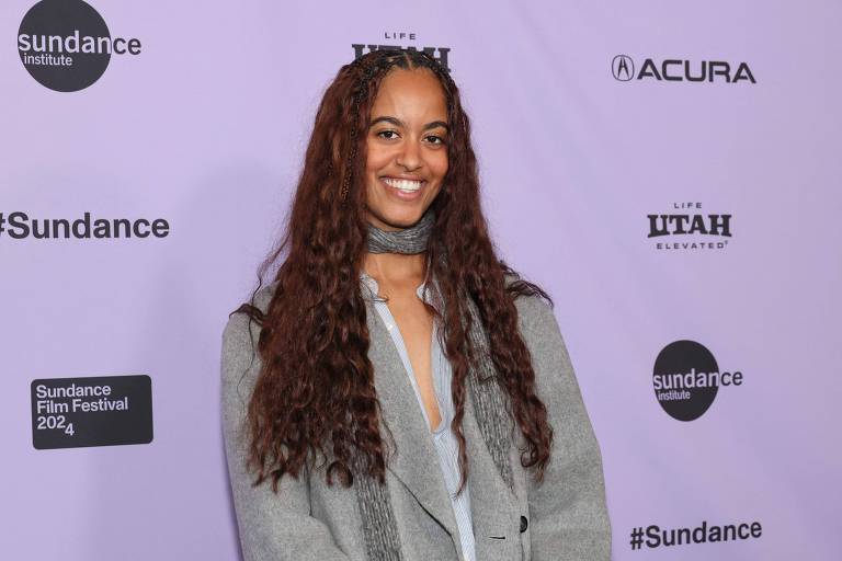 Uma mulher com cabelo cacheado e longo, usando uma blusa clara e um casaco cinza, está sorrindo em um evento. O fundo é roxo com logotipos do Sundance Institute e Acura, além de texto que diz 'Sundance Film Festival 2024'.