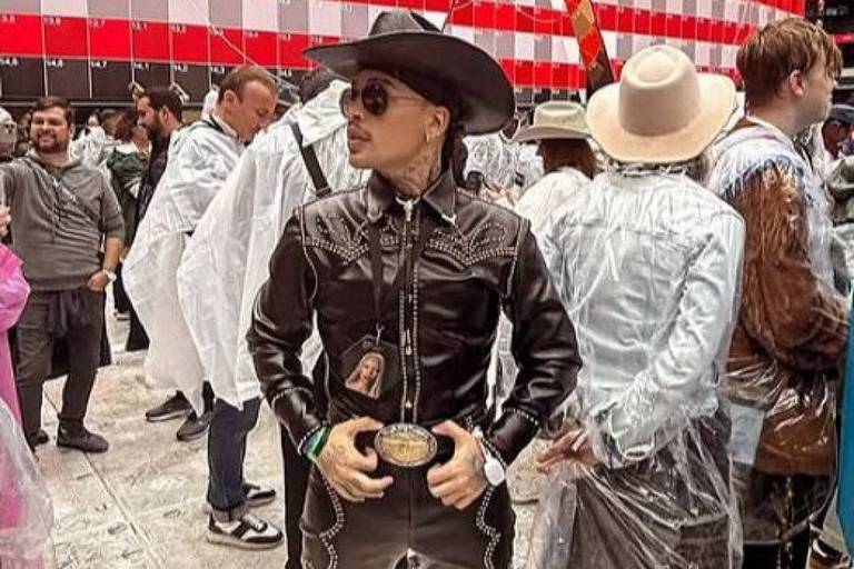 A imagem mostra uma pessoa vestindo um traje de cowboy, que inclui uma camisa preta com detalhes em branco, calças escuras e um chapéu de cowboy. A pessoa está em uma pose confiante, com as mãos na cintura. Ao fundo, há várias outras pessoas, algumas usando capas de chuva, e um grande painel com listras vermelhas e brancas. O ambiente parece ser um evento ao ar livre.