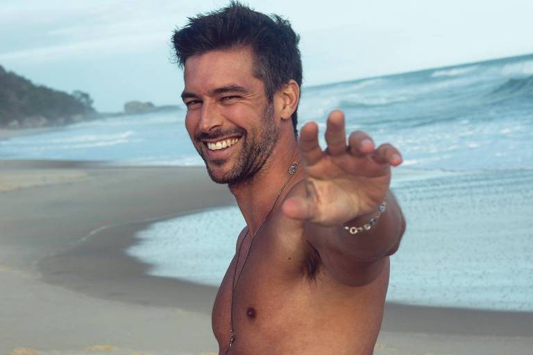 Um homem sorridente está posando em uma praia. Ele está sem camisa, mostrando um corpo atlético, e usa jeans. O homem está de lado, com um braço estendido em direção à câmera, enquanto o mar e a areia estão ao fundo. O céu está claro e há algumas nuvens.