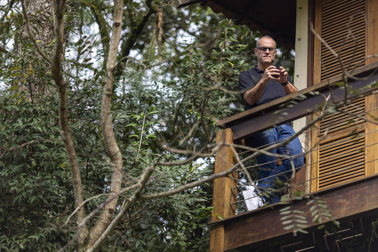 Um homem está em uma varanda de uma casa na árvore, segurando um objeto com as duas mãos. Ele usa uma camiseta preta e jeans. Ao redor, há árvores e folhagens, criando um ambiente natural. A varanda é de madeira e possui uma grade. O homem parece estar observando algo à sua frente.