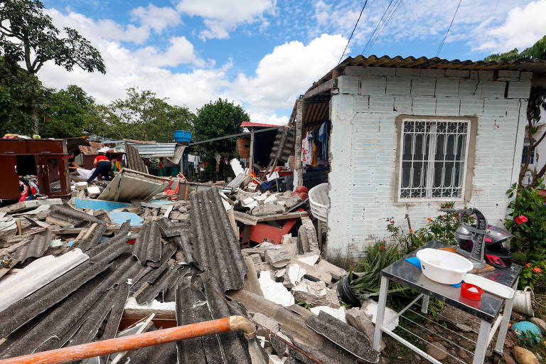 A imagem mostra uma área urbana devastada, com destroços de telhados e materiais de construção espalhados pelo chão. Há uma casa de paredes claras e uma janela com grades visíveis. Ao fundo, outras estruturas danificadas são vistas, e algumas pessoas estão trabalhando na limpeza dos escombros. O céu está parcialmente nublado, e há vegetação ao redor.