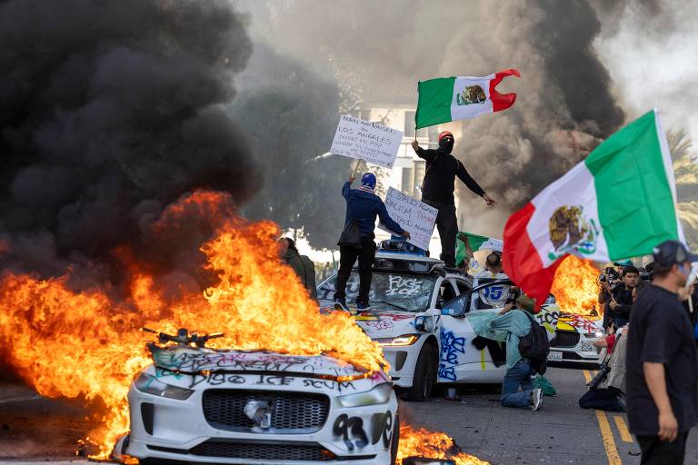 A imagem mostra uma cena de protesto com veículos em chamas e fumaça preta se elevando. Algumas pessoas estão segurando bandeiras do México, enquanto outras estão em pé sobre os carros. Há cartazes visíveis, e o ambiente parece caótico devido ao fogo e à fumaça.