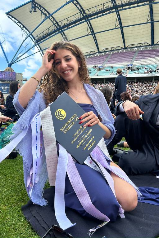 Uma jovem mulher está sentada em um campo, segurando um diploma. Ela tem cabelos cacheados e está vestindo um vestido azul, com uma estola ou xale claro. Ao fundo, há outras pessoas em trajes de formatura e uma estrutura de estádio visível. O céu está parcialmente nublado.