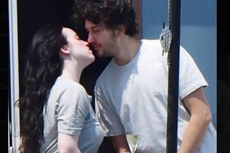A imagem mostra um casal se beijando em uma varanda. Ambos estão usando camisetas de cores claras. A mulher tem cabelo longo e escuro, enquanto o homem tem cabelo castanho e um pouco encaracolado. Ao lado deles, há uma taça de champanhe sobre a mesa. O fundo é de uma parede clara e a cena parece ser iluminada pelo sol.
