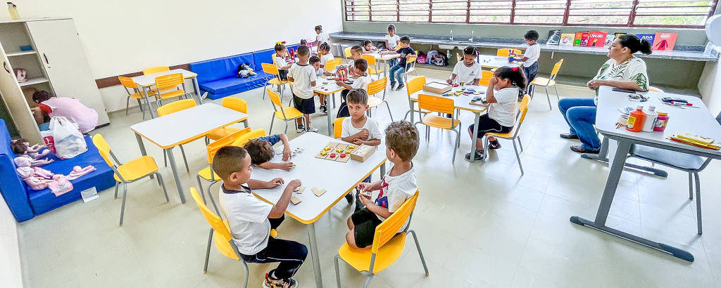 A imagem mostra uma sala de aula com várias crianças sentadas em mesas. As crianças estão vestidas com camisetas brancas e algumas estão comendo. Há mesas brancas e cadeiras amarelas. Ao fundo, há uma parede com janelas grandes que permitem a entrada de luz natural. Uma professora ou cuidadora está sentada em uma mesa ao fundo, observando as crianças. O ambiente é bem iluminado e organizado.