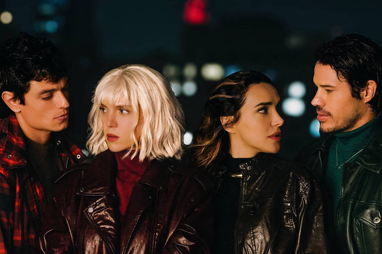 A imagem mostra quatro pessoas em um ambiente urbano à noite. Duas delas estão à esquerda, um homem com cabelo escuro e uma mulher com cabelo loiro curto. À direita, há uma mulher com cabelo castanho e uma expressão séria, e um homem com cabelo escuro e barba. Todos estão vestidos com jaquetas escuras e parecem estar em uma conversa intensa, com luzes da cidade ao fundo.