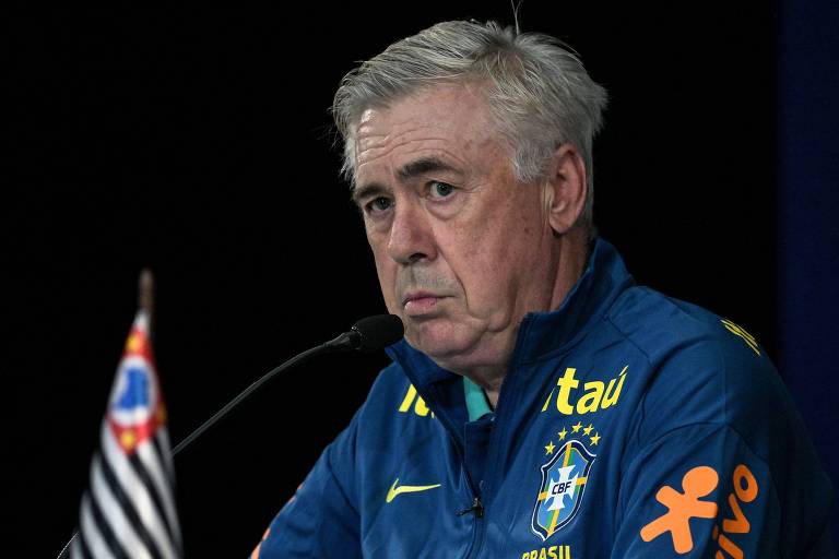 Um homem de cabelo grisalho, vestindo um agasalho azul com detalhes em amarelo e o escudo da seleção brasileira de futebol, está sentado em uma mesa de coletiva de imprensa. Ele parece sério e está olhando para frente. À sua esquerda, há uma bandeira do São Paulo. O fundo é escuro, destacando a figura do homem.