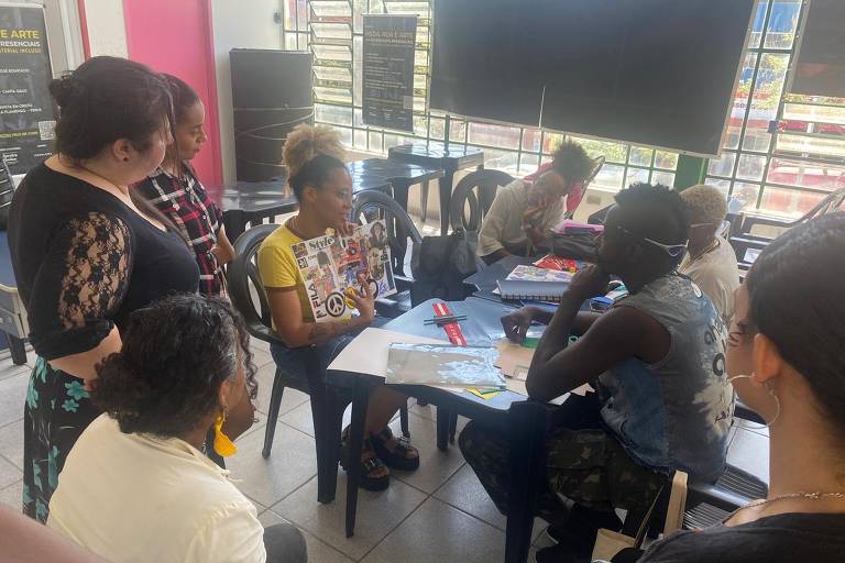 A imagem mostra um grupo de pessoas em um ambiente interno, participando de uma atividade criativa. Uma mulher com cabelo cacheado e blusa amarela está segurando uma revista e conversando com outras pessoas que a observam atentamente. Há uma mesa com materiais de arte, como papéis e tesouras. Ao fundo, é possível ver janelas grandes que permitem a entrada de luz natural. O ambiente parece ser um espaço comunitário ou de workshop.