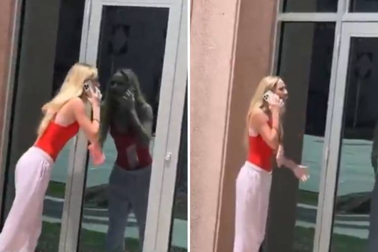 A imagem mostra uma mulher com cabelo loiro longo, vestindo uma blusa vermelha e calças brancas, falando ao telefone. Ela está em frente a uma porta de vidro, onde seu reflexo é visível. A mulher parece estar gesticulando enquanto conversa, e o ambiente ao fundo é iluminado, sugerindo que é um dia ensolarado.