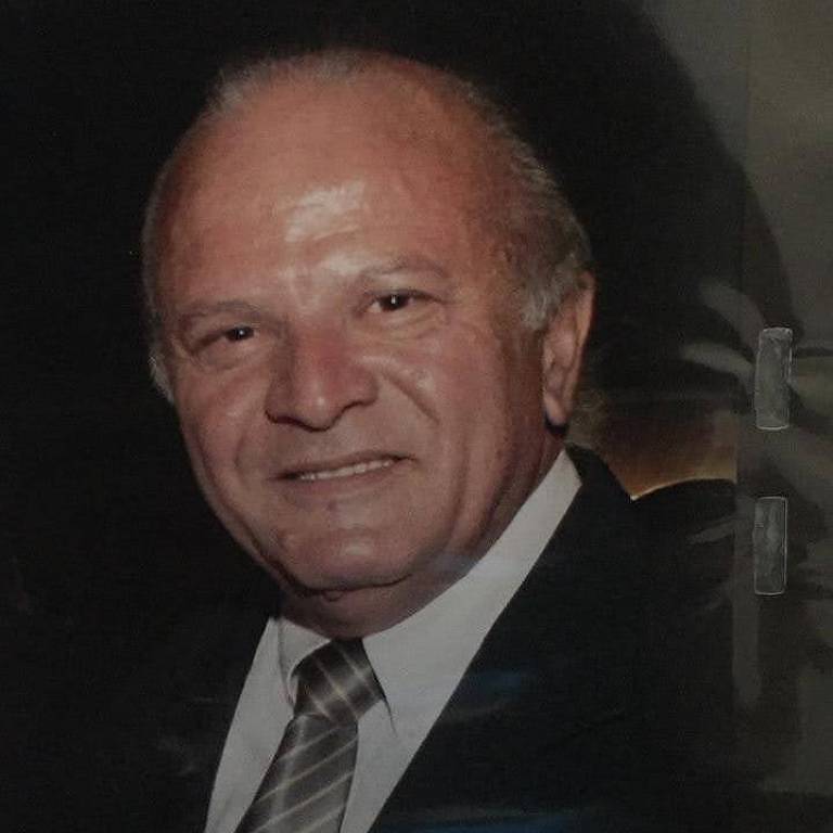 A imagem mostra um homem sorridente, com cabelo curto e grisalho, usando um terno escuro e uma gravata listrada. Ele está posicionado de lado, olhando para a câmera, com um fundo desfocado que sugere um ambiente formal.