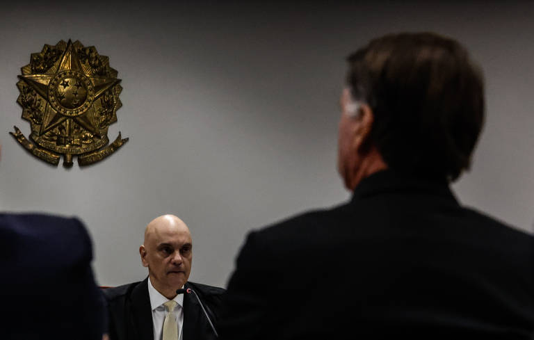A imagem mostra uma audiência onde um homem careca, vestido com um terno escuro e gravata clara, está sentado à mesa, olhando para frente. Ao fundo, há um emblema dourado na parede. Na frente dele, são visíveis as costas de dois homens, um deles com cabelo castanho e liso, vestido de preto. A cena sugere um ambiente formal, possivelmente uma sessão de tribunal ou uma reunião de autoridades.