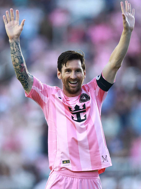 Um jogador de futebol está em campo, vestindo uma camisa rosa com listras verticais. Lionel Messi tem os braços levantados e está sorrindo, demonstrando alegria. O fundo é desfocado, mas há uma multidão visível, com muitas pessoas vestindo roupas em tons de rosa.