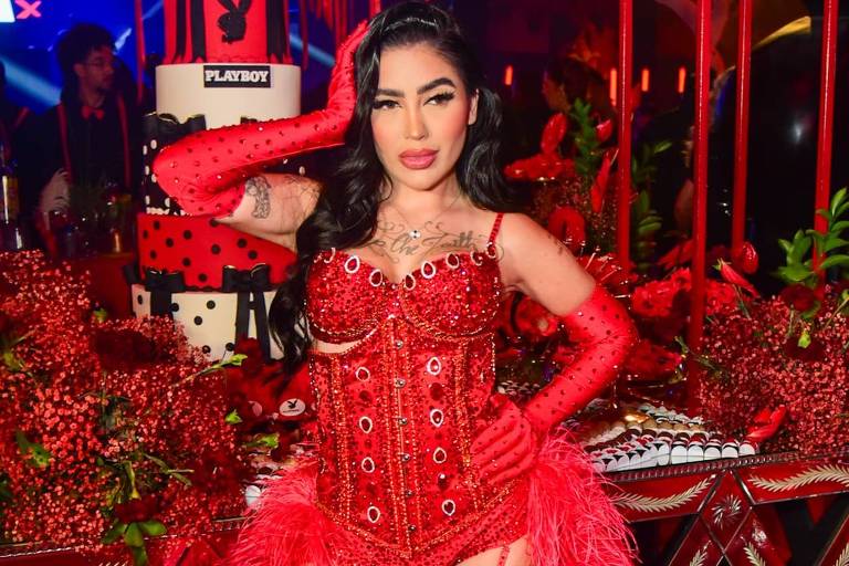 A imagem mostra uma mulher em um evento festivo, vestindo um traje vermelho brilhante que inclui um corpete e meias altas. Ela está posando com uma mão na cabeça e a outra na cintura, cercada por uma decoração exuberante em tons de vermelho, incluindo um bolo grande ao fundo. O ambiente é iluminado e festivo, com várias mesas decoradas.