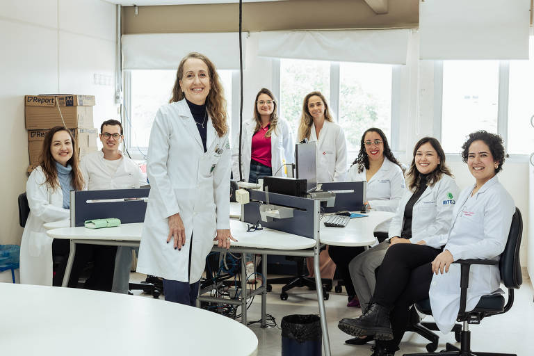 A imagem mostra um grupo de oito profissionais de saúde, todos vestidos com jalecos brancos, em um ambiente de escritório. No centro, uma mulher mais velha está em pé, sorrindo, enquanto os outros estão sentados ao redor de mesas com computadores. O ambiente é bem iluminado, com janelas ao fundo e caixas empilhadas em um canto.