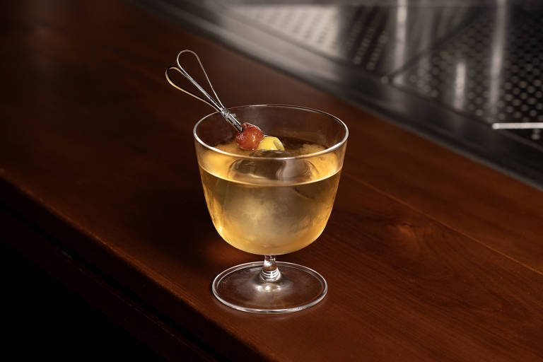 Um copo de coquetel transparente com uma bebida amarelada, decorado com uma cereja e uma fatia de limão presa a um palito. O copo está sobre uma superfície de madeira escura, e ao fundo é possível ver uma superfície metálica com pequenos furos.