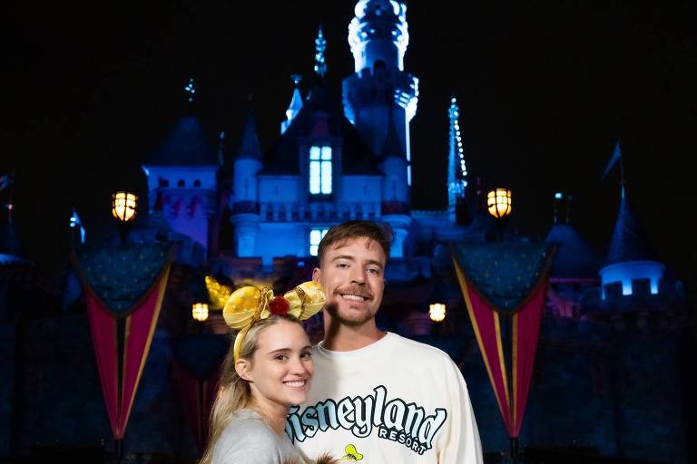 Um casal posando em frente ao castelo iluminado do Disneyland à noite. A mulher usa orelhas de Mickey e segura dois brinquedos de pelúcia, enquanto o homem está vestido com um suéter que diz 'Disneyland'. O castelo ao fundo está iluminado em tons de azul e roxo, criando uma atmosfera mágica.