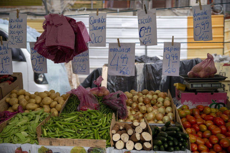 A imagem mostra uma barraca de feira com diversos tipos de frutas e verduras expostas. Há batatas, cebolas, pimentões, tomates, e outros vegetais. Os preços estão visíveis em placas de papel penduradas, com valores como 2,00, 4,99 e 9,99. O ambiente parece ser ao ar livre, com um fundo desfocado de um veículo e uma estrutura de barraca.