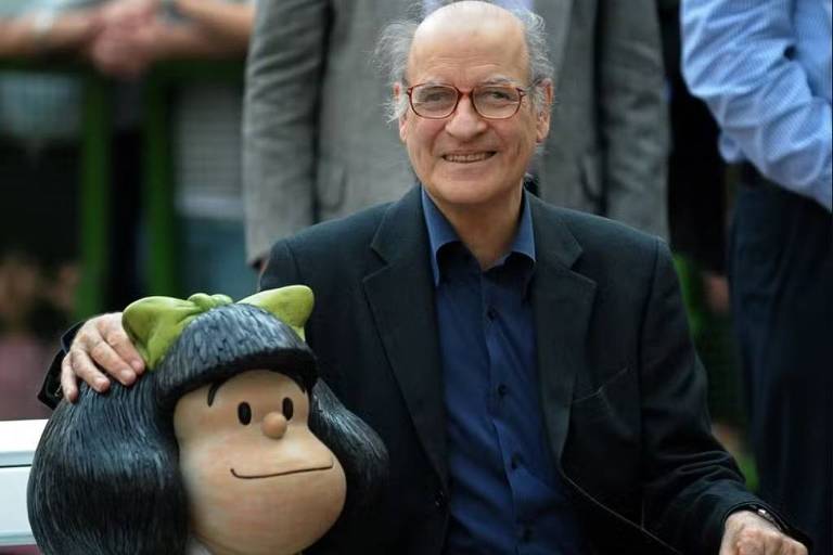 Um homem de óculos e cabelo grisalho está sentado ao lado de uma estátua da personagem Mafalda, que tem cabelo preto e usa um vestido verde. O homem está sorrindo e tem a mão apoiada na cabeça da estátua. Ao fundo, há pessoas em pé, mas não são claramente visíveis.