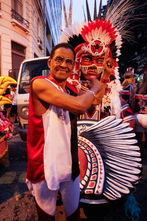 A imagem mostra um homem sorridente em um traje de carnaval, posando com um dedo apontado para cima. Ele está ao lado de uma figura vestida com uma fantasia elaborada, que inclui penas e pintura facial. O fundo apresenta um veículo e outras pessoas, sugerindo um ambiente festivo de carnaval.