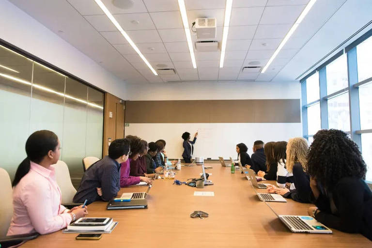 A imagem mostra uma sala de conferência com uma longa mesa de madeira. Várias pessoas estão sentadas ao redor da mesa, todas atentas a uma apresentação que está sendo feita por uma mulher que está de pé em frente a um quadro branco. A sala possui janelas grandes que permitem a entrada de luz natural. A maioria das pessoas está usando laptops e algumas têm garrafas de água e cadernos na mesa.