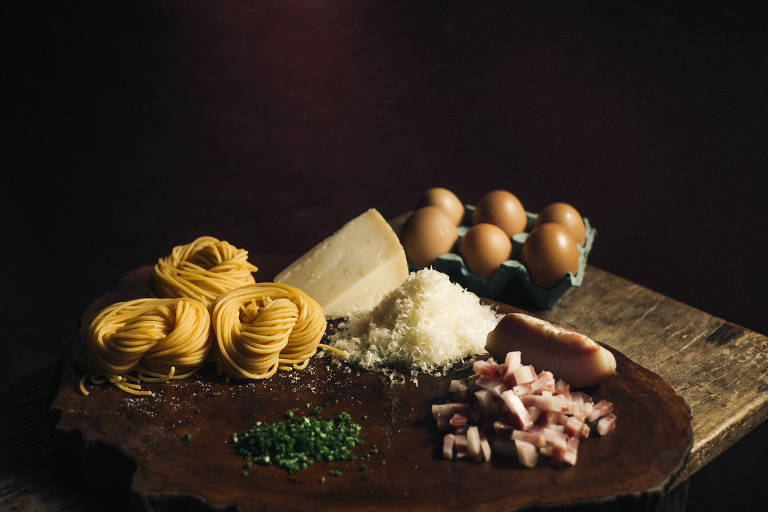 A imagem mostra uma tábua de madeira com diversos ingredientes dispostos. Há um ninho de macarrão amarelo, um pedaço de queijo parmesão, ovos em uma cartela, queijo ralado, cebolinha picada e pedaços de bacon. O fundo é escuro, destacando os ingredientes.