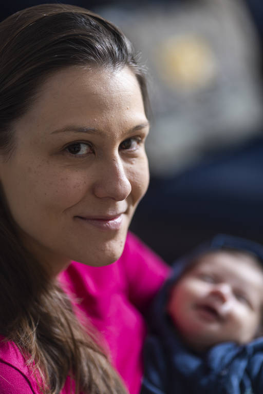 A imagem mostra uma mulher sorrindo, com cabelo longo e castanho, usando uma blusa rosa. Ela está segurando um bebê que parece estar dormindo em seu colo, vestido com um macacão azul escuro. Ao fundo, há um sofá com uma almofada que não é claramente visível.