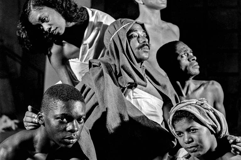 A imagem em preto e branco mostra um grupo de seis pessoas em poses dramáticas. No centro, uma figura masculina vestida com um manto escuro, cercada por outras cinco pessoas, algumas com expressões sérias e outras em poses contemplativas. O fundo é desfocado, destacando os rostos e as expressões dos indivíduos. A iluminação cria um contraste forte, acentuando as sombras e as texturas das roupas e da pele.