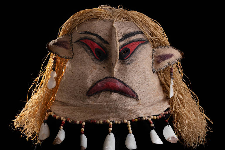 A imagem mostra uma máscara com características faciais expressivas, incluindo olhos vermelhos e uma boca com uma expressão de descontentamento. A máscara é adornada com cabelo loiro e possui orelhas, além de colares com contas e conchas na parte inferior. O fundo da imagem é preto, destacando a máscara.