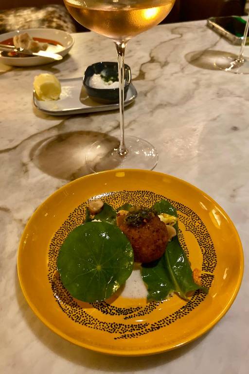A imagem mostra um prato gourmet em um fundo de mesa de mármore. O prato é amarelo e contém uma bolinho frito, folhas verdes e um molho branco. Ao fundo, há uma taça de vinho e outros pratos pequenos em uma mesa bem decorada.