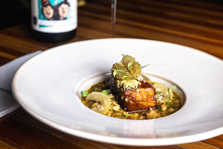 A imagem mostra um prato gourmet com um pedaço de carne grelhada, decorado com folhas verdes, servido sobre uma cama de legumes. Ao lado, há uma taça de vinho tinto e uma garrafa de vinho com um rótulo que apresenta três pessoas. O fundo é desfocado, com plantas visíveis.