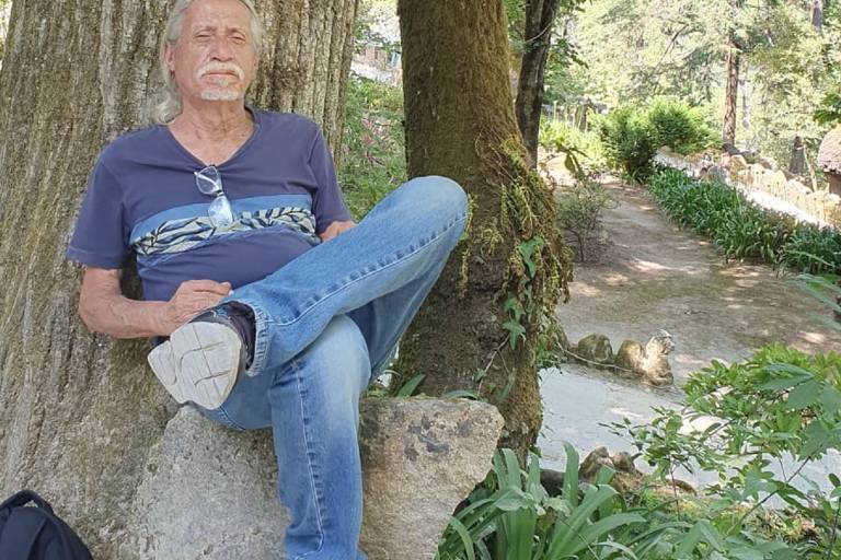 Um homem está sentado em um tronco de árvore, com uma perna cruzada sobre a outra. Ele usa uma camiseta azul e jeans. Ao fundo, há um caminho cercado por vegetação e árvores. A luz do sol ilumina a cena, criando um ambiente natural e tranquilo.