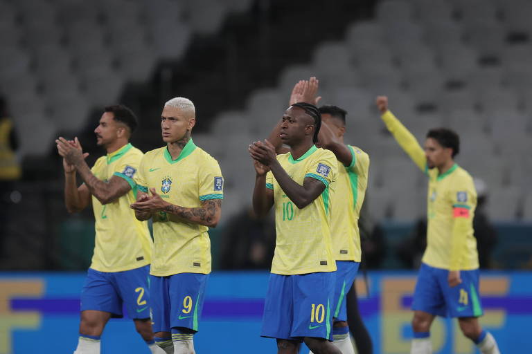 Um grupo de jogadores da seleção brasileira de futebol, vestidos com uniformes amarelos e azuis, está aplaudindo. Eles estão em um estádio, com algumas pessoas ao fundo. O jogador no centro, com cabelo platinado, é o mais destacado, enquanto outros jogadores estão em posições variadas, mostrando expressões de gratidão ou celebração.