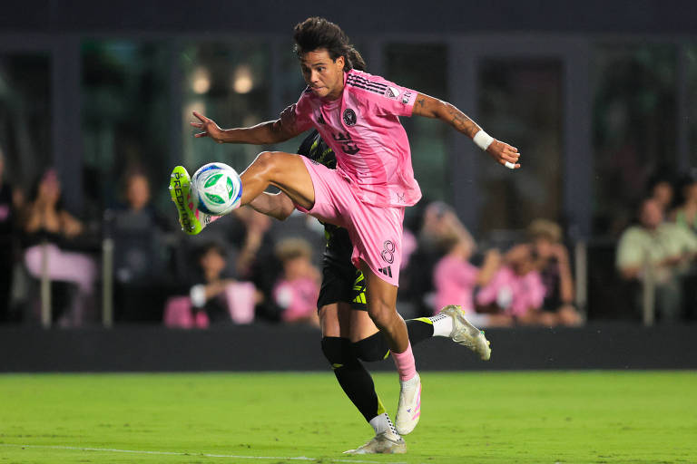 Um jogador de futebol está em ação, vestindo um uniforme rosa. Telasco Segovia está levantando uma perna enquanto tenta controlar a bola, que está próxima a ele. O fundo mostra uma multidão assistindo ao jogo, com algumas pessoas vestindo camisetas em tons de rosa. O campo de futebol é bem iluminado, e a atmosfera parece ser de um evento esportivo animado.