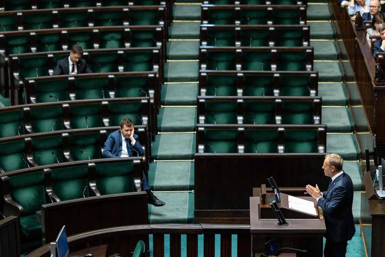 A imagem mostra um ambiente de uma sessão legislativa com várias cadeiras vazias. Um homem está sentado em uma das cadeiras, enquanto outro homem, que está de pé, fala em um púlpito. O ambiente é predominantemente em tons de verde e madeira escura, com um layout que sugere um espaço formal de debate.