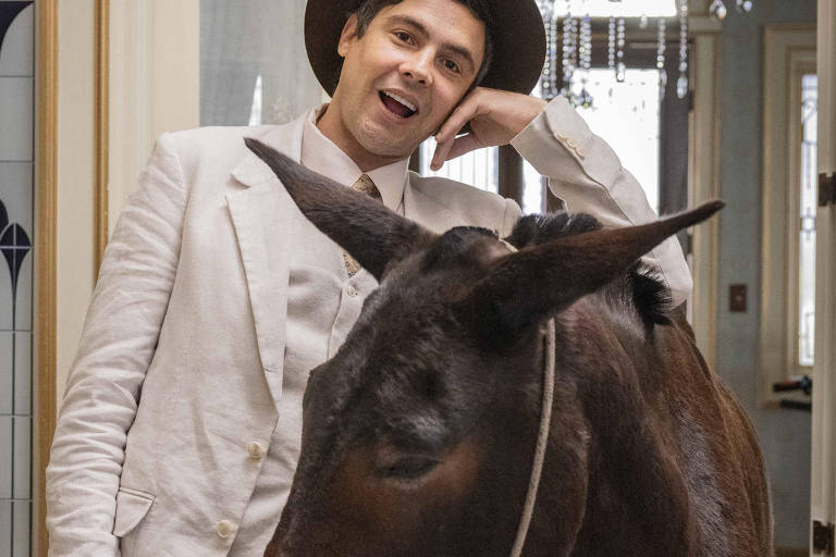 Um homem vestido com um terno claro e chapéu está posando ao lado de um burro em um ambiente interno. O homem sorri e parece estar se divertindo, enquanto o burro está com a cabeça baixa, próximo ao homem. O fundo apresenta uma decoração elegante, com um lustre visível e paredes decoradas.