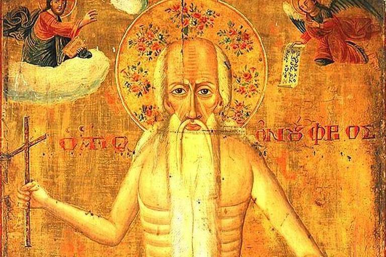 A imagem apresenta um ícone religioso de um santo, representado como uma figura alta e esquelética, com longos cabelos e barba. Ele segura um bastão em uma mão e uma tábua com inscrições na outra. A parte inferior do corpo é coberta por folhas verdes, e ao fundo há uma paisagem com árvores e montanhas. No topo, há figuras de anjos e nuvens.
