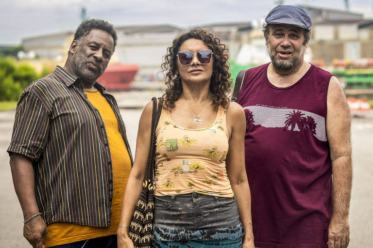 Três pessoas estão em pé em um ambiente urbano, possivelmente próximo a um porto. À esquerda, um homem de pele escura, com cabelo curto e barba, usa uma camisa listrada e calças escuras. No centro, uma mulher de pele clara, com cabelo cacheado e óculos escuros, veste uma blusa amarela com estampas florais e uma saia longa. À direita, um homem de pele clara, com cabelo curto e barba, usa uma camiseta roxa e calças brancas. Ao fundo, há estruturas industriais e equipamentos de porto.