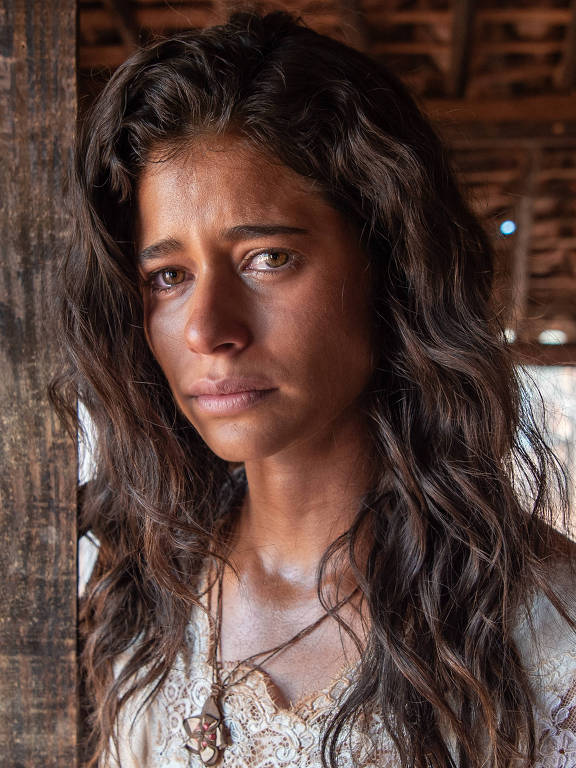A imagem mostra uma mulher com cabelo longo e ondulado, olhando para a câmera com uma expressão séria. Ela está vestindo uma blusa clara com detalhes em renda e está posicionada ao lado de uma parede de madeira escura. O ambiente ao fundo parece rústico, com uma estrutura de madeira visível e iluminação suave.