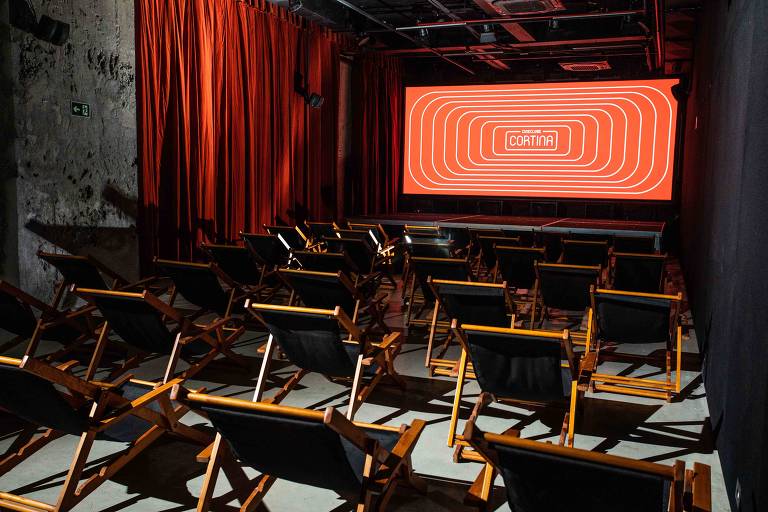 A imagem mostra uma sala de cinema com cadeiras de madeira dobráveis dispostas em fileiras. Na frente, há uma tela grande com um fundo vermelho e um design gráfico em linhas onduladas. As paredes são de cor escura e há cortinas vermelhas ao fundo, criando um ambiente aconchegante e moderno.