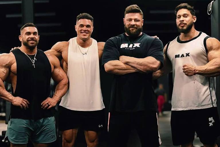 Quatro homens musculosos estão posando juntos em um ambiente de academia. Eles estão vestidos com roupas de treino, com destaque para camisetas sem mangas e shorts. O fundo é escuro, com equipamentos de musculação visíveis. Todos os homens têm expressões confiantes e estão em pé, mostrando seus músculos.