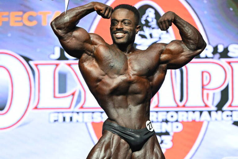 Um fisiculturista em pose de competição, exibindo músculos bem definidos. Ele está em um palco com um fundo colorido que inclui o logotipo do evento 'Olympia'. O fisiculturista usa um traje de competição preto e está em uma pose de flexão, mostrando seus braços e peito musculosos. O ambiente parece ser uma competição de fisiculturismo, com iluminação focada no competidor.