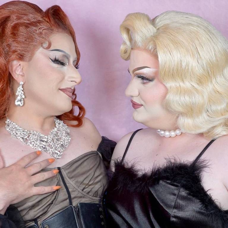 A imagem mostra duas drag queens em um fundo rosa. A primeira drag, à esquerda, tem cabelo ruivo e usa um vestido cinza escuro com um corpete, além de um colar brilhante. A segunda drag, à direita, tem cabelo loiro platinado e usa um vestido preto com detalhes de penas. Ambas estão se olhando com expressões amigáveis e sorridentes.