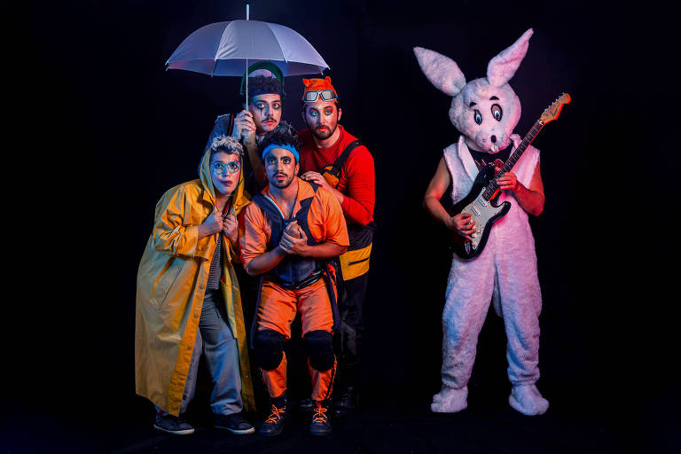 A imagem mostra um grupo de cinco pessoas em trajes coloridos e fantasias. Quatro delas estão em pé, com expressões faciais divertidas, enquanto uma delas segura um guarda-chuva. A quinta pessoa, à direita, está vestida como um coelho e toca guitarra. O fundo é escuro, destacando as cores vibrantes das roupas e a iluminação do grupo.