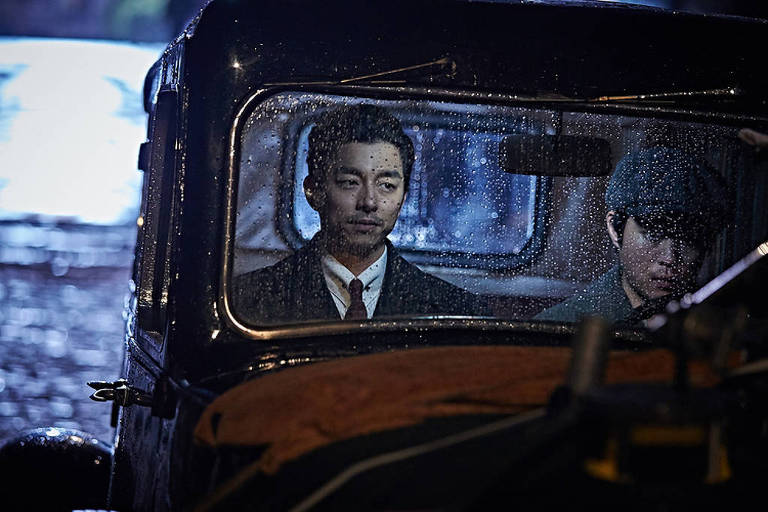 A imagem mostra dois homens dentro de um carro antigo, com a chuva caindo sobre o vidro. Um dos homens está usando um terno escuro e parece pensativo, enquanto o outro, ao lado, está com um chapéu e uma expressão mais séria. O ambiente externo é pouco iluminado, sugerindo um clima noturno e melancólico.