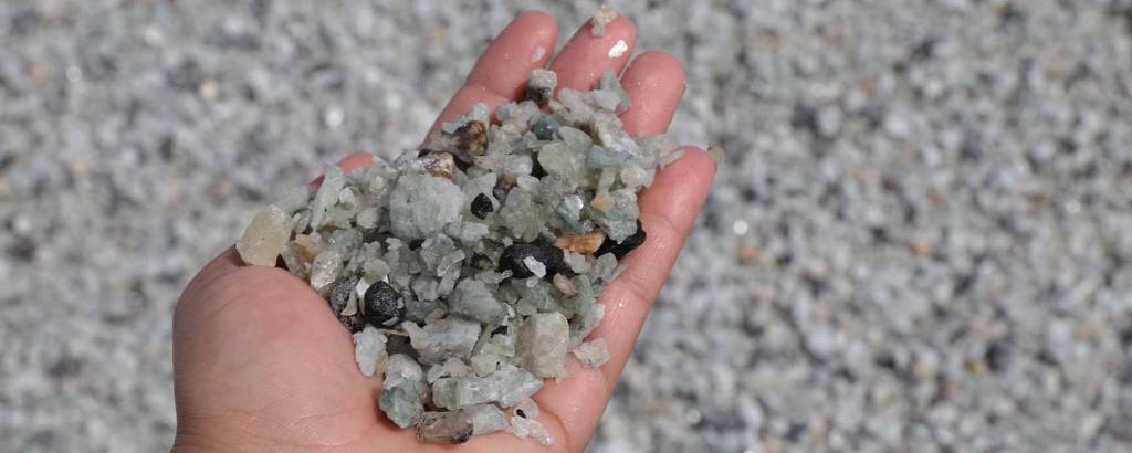 Uma mão humana segurando uma quantidade de pedras e cristais de diferentes tamanhos e cores, incluindo tons de verde, cinza e preto. Ao fundo, há uma superfície de pedras menores, que parece ser uma praia ou um local com cascalho.
