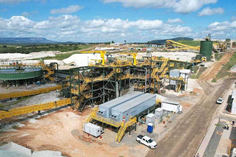 A imagem mostra uma grande instalação industrial, possivelmente uma planta de mineração ou processamento. Há várias estruturas metálicas, incluindo correias transportadoras e tanques, além de caminhões e equipamentos pesados. O céu está parcialmente nublado e a área ao redor é predominantemente rural, com vegetação visível ao fundo.