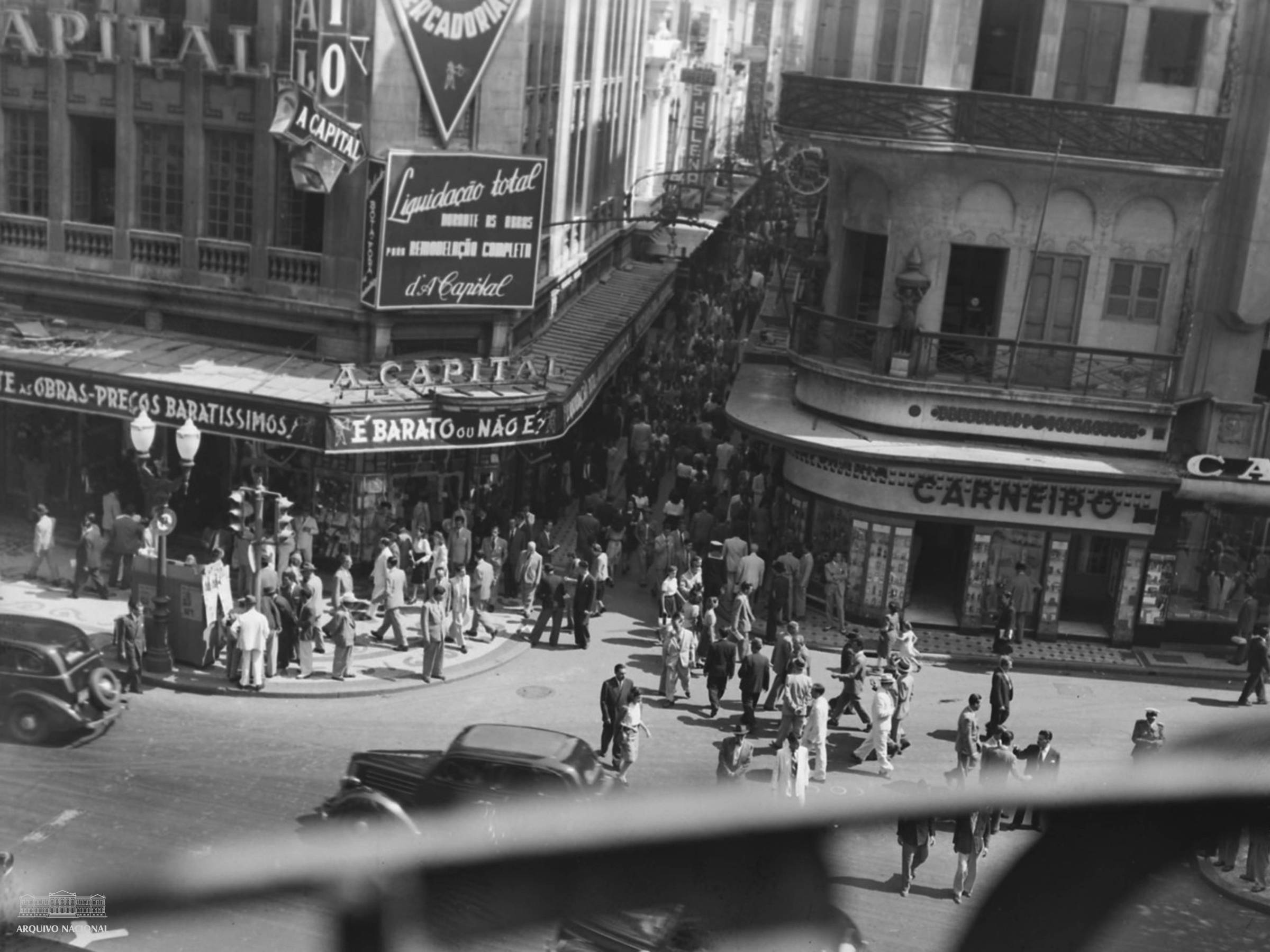 A imagem mostra uma cena movimentada da Rua do Ouvidor, na cidade do Rio de Janeiro, capturada em preto e branco. Há muitas pessoas caminhando nas calçadas e na rua, com alguns carros antigos visíveis. Várias lojas estão presentes, incluindo uma com o nome 'A Capital' e outra chamada 'Carneiro'. Placas e anúncios estão visíveis, incluindo um que diz 'Liquidação total'. O ambiente parece ser de uma área comercial movimentada.
