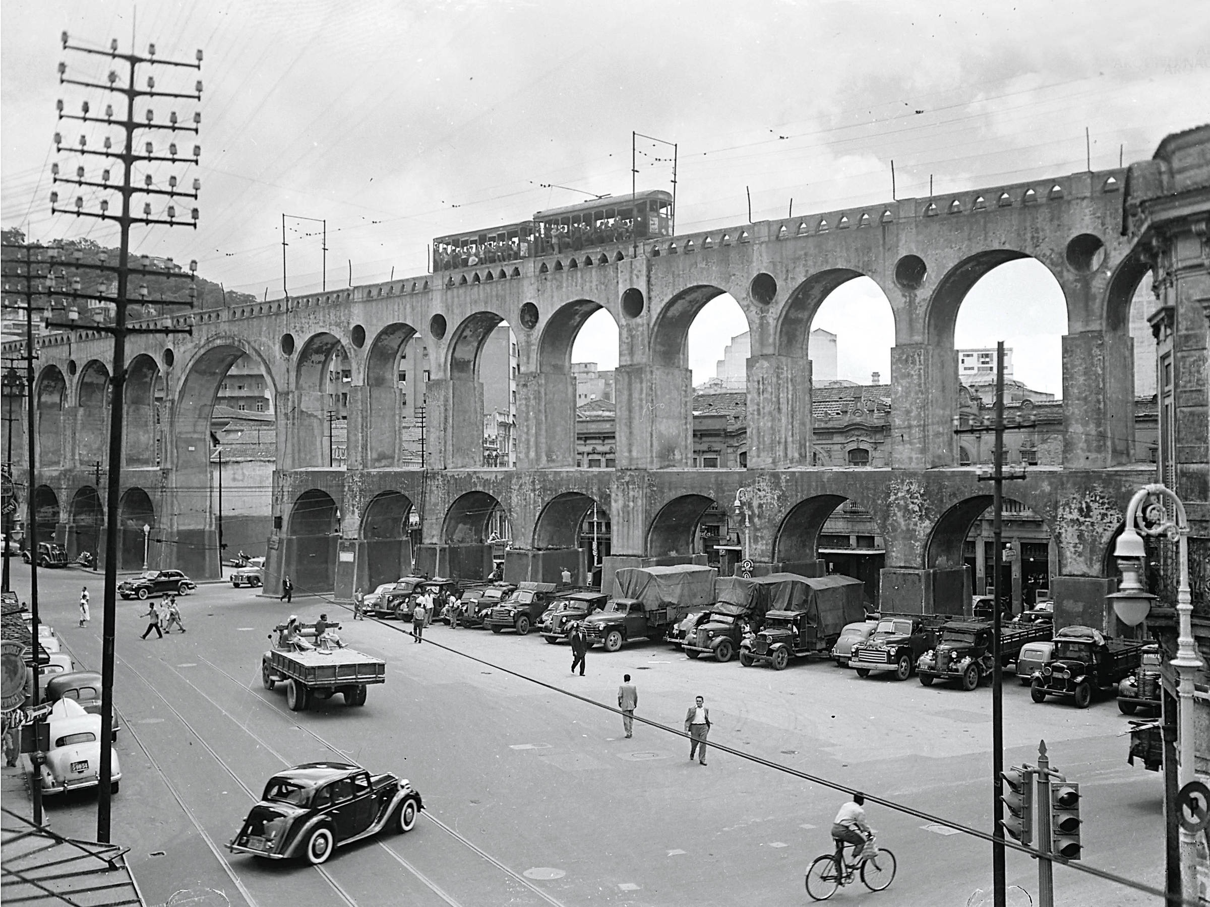 A imagem mostra um viaduto com arcos, que se eleva sobre uma rua movimentada. Abaixo do viaduto, há vários carros estacionados e em movimento, incluindo um caminhão e um carro de passeio. Algumas pessoas estão atravessando a rua, enquanto um ciclista passa. Ao fundo, há edifícios e postes de eletricidade. O céu está nublado, e a imagem é em preto e branco, sugerindo uma época passada.